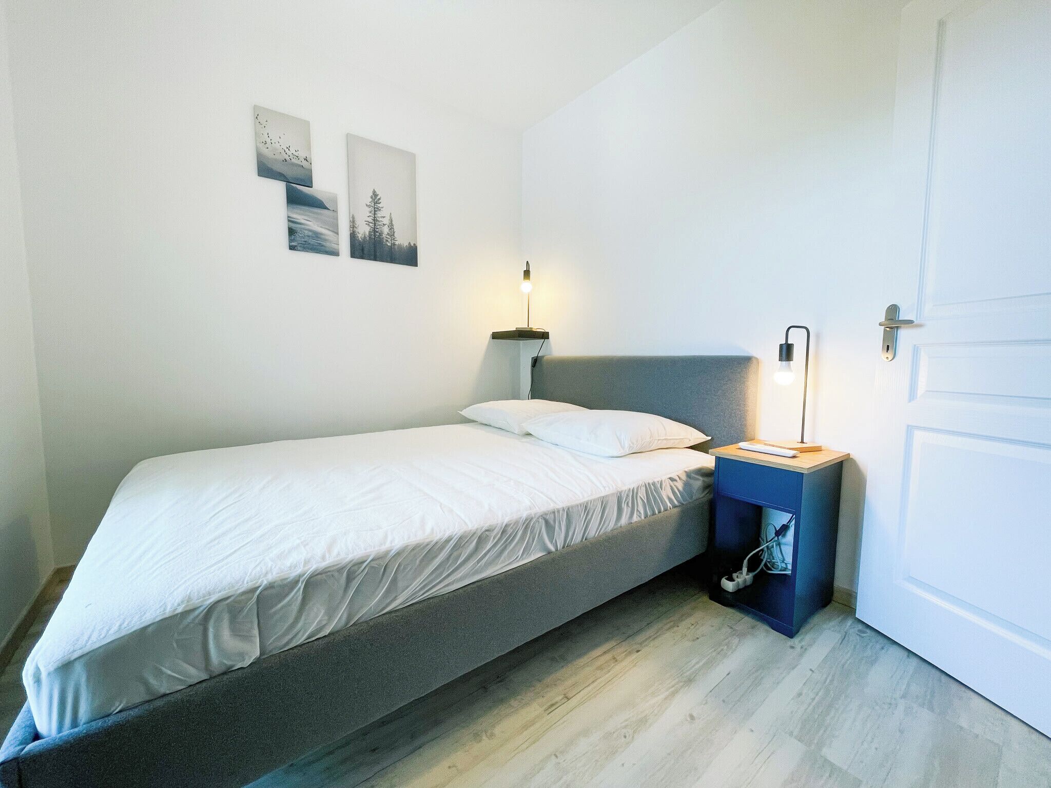A single bed with a grey headboard in a room with white walls. There are two wall-mounted lamps on either side of the bed.