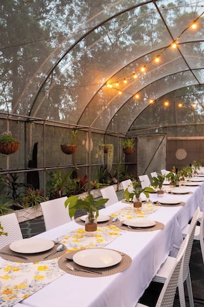 A greenhouse with tables set for dining, surrounded by hanging planters and string lights.