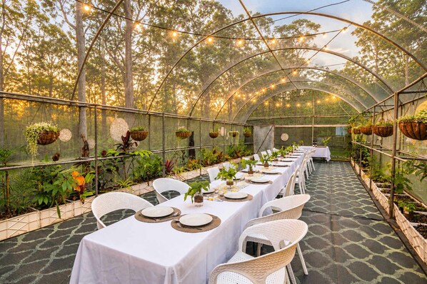 Une serre aménagée avec des tables pour les repas, entourée de plantes et de suspensions florales.