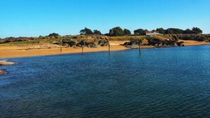 Un paisaje costero con una playa arenosa, agua tranquila y un edificio blanco de techo rojo a lo lejos.