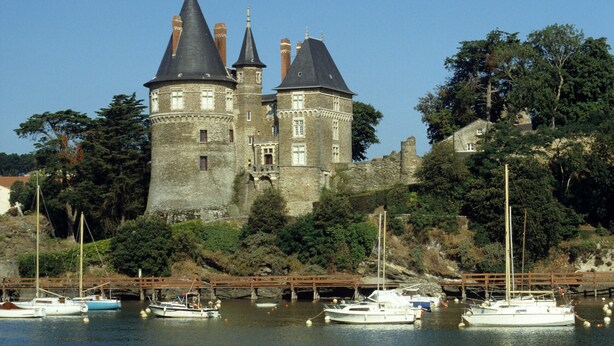 Un castillo histórico con múltiples torres, ubicado a orillas de un río tranquilo donde hay varias embarcaciones amarradas.