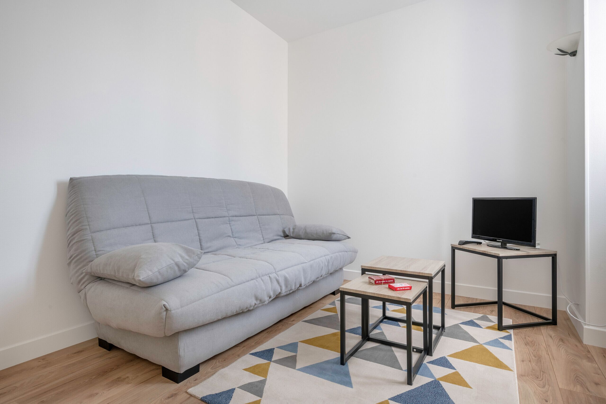 A modern living room with a grey sofa, a small wooden coffee table, a TV on a stand, and a patterned rug.
