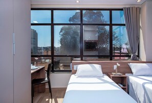 Ein modernes Hotelzimmer mit großem Fenster, einem Schreibtisch, zwei Betten und Blick auf die Stadt.