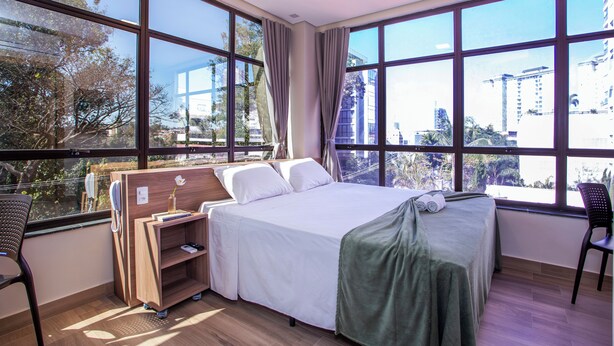 Habitación de hotel con una cama grande, un escritorio y una silla. Amplias ventanas ofrecen vistas a edificios y árboles.
