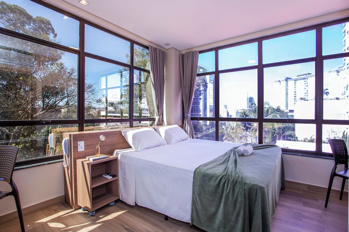 Ein Hotelzimmer mit einem großen Bett, einem Schreibtisch und einem Stuhl. Große Fenster bieten Blick auf Gebäude und Bäume.