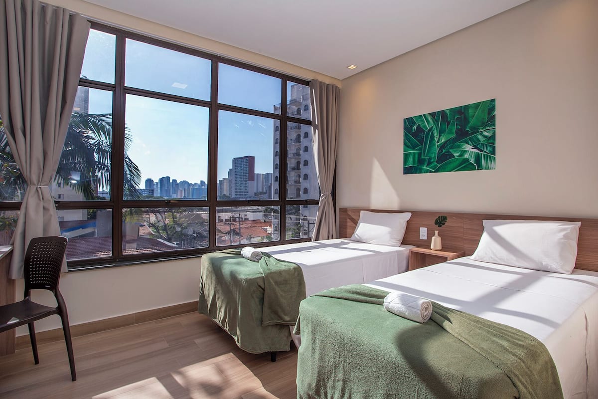 Ein Hotelzimmer mit zwei Betten, Blick auf die Stadtlandschaft, einer Zimmerpflanze und einem Sessel.