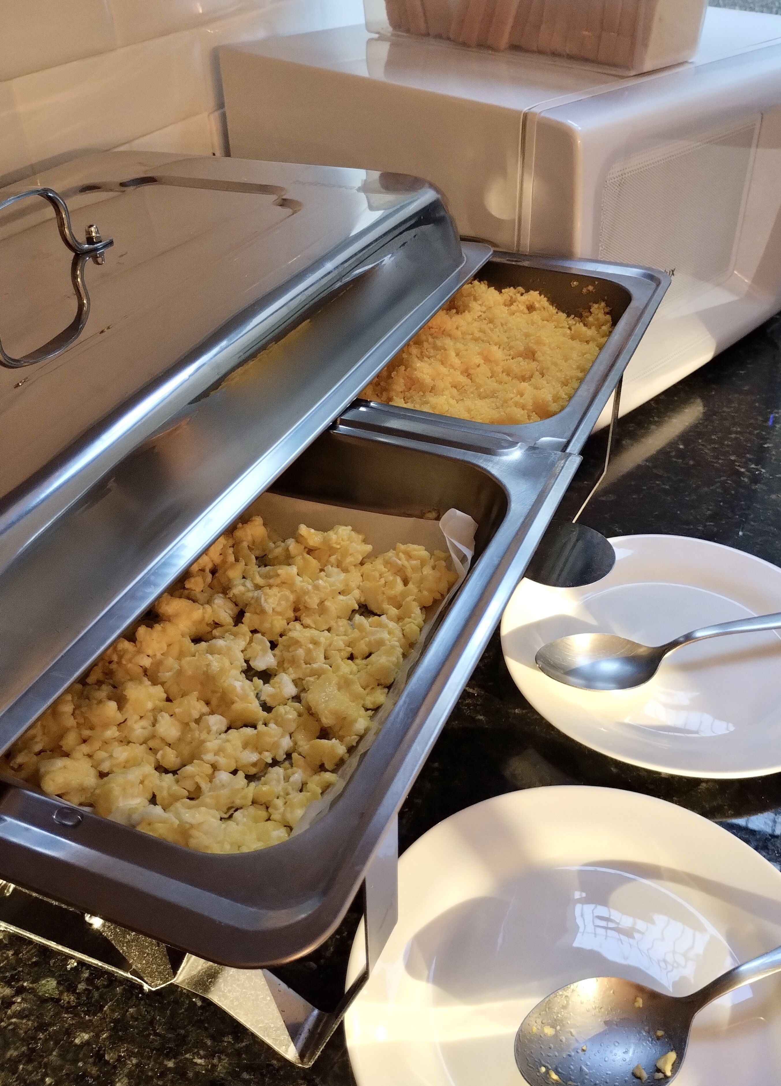 A buffet setup with two trays of food, a spoon, and a plate on a countertop.