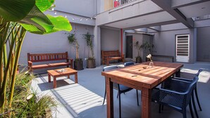 Un patio al aire libre con mesa y bancos de madera, rodeado de plantas en macetas y una planta de plátano grande.