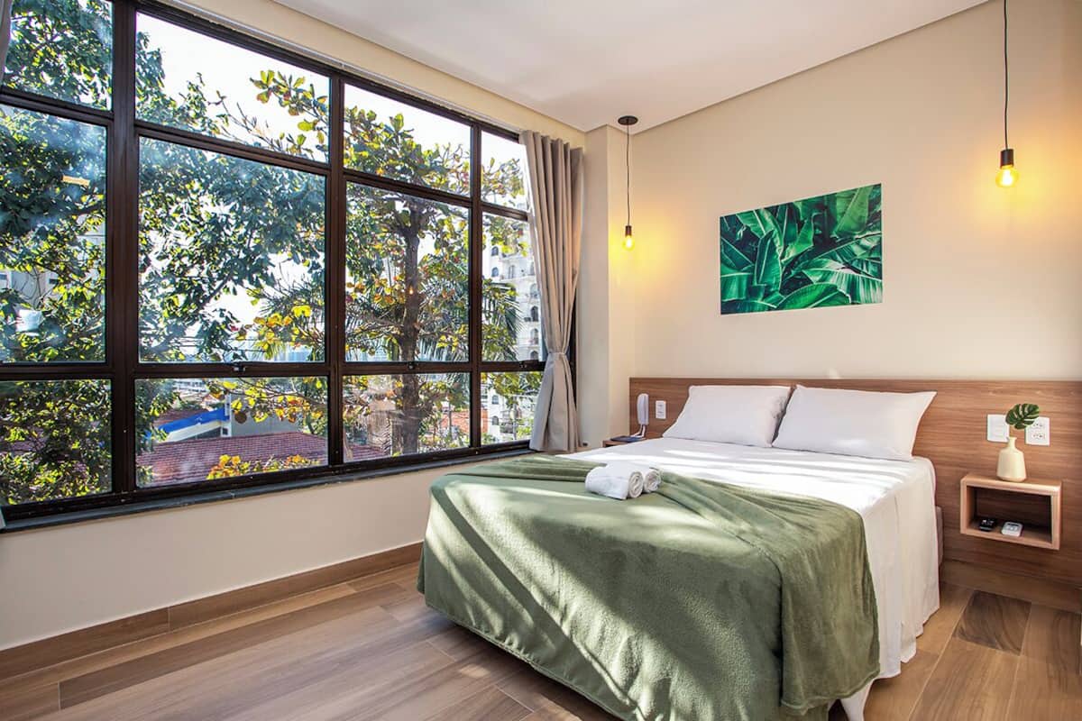 Ein Hotelzimmer mit großem Fenster, einem Bett mit grüner Decke, einem Nachttisch mit Pflanze und einer wandmontierten Lampe.