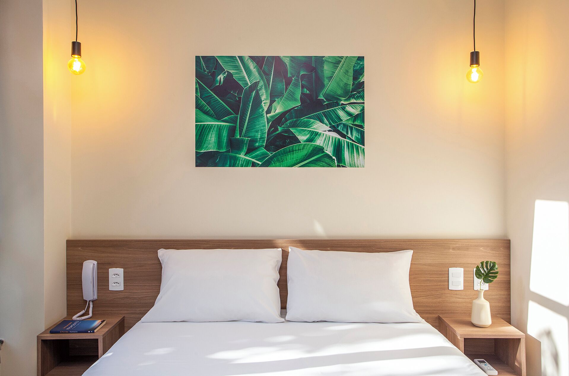 A hotel room with a wooden headboard, two white pillows, a bed, a nightstand with a phone and a book, a wall-mounted light fixture, and a wall art of green leaves.