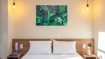 A hotel room with a wooden headboard, two white pillows, a bed, a nightstand with a phone and a book, a wall-mounted light fixture, and a wall art of green leaves.