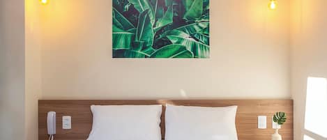 A hotel room with a wooden headboard, two white pillows, a bed, a nightstand with a phone and a book, a wall-mounted light fixture, and a wall art of green leaves.