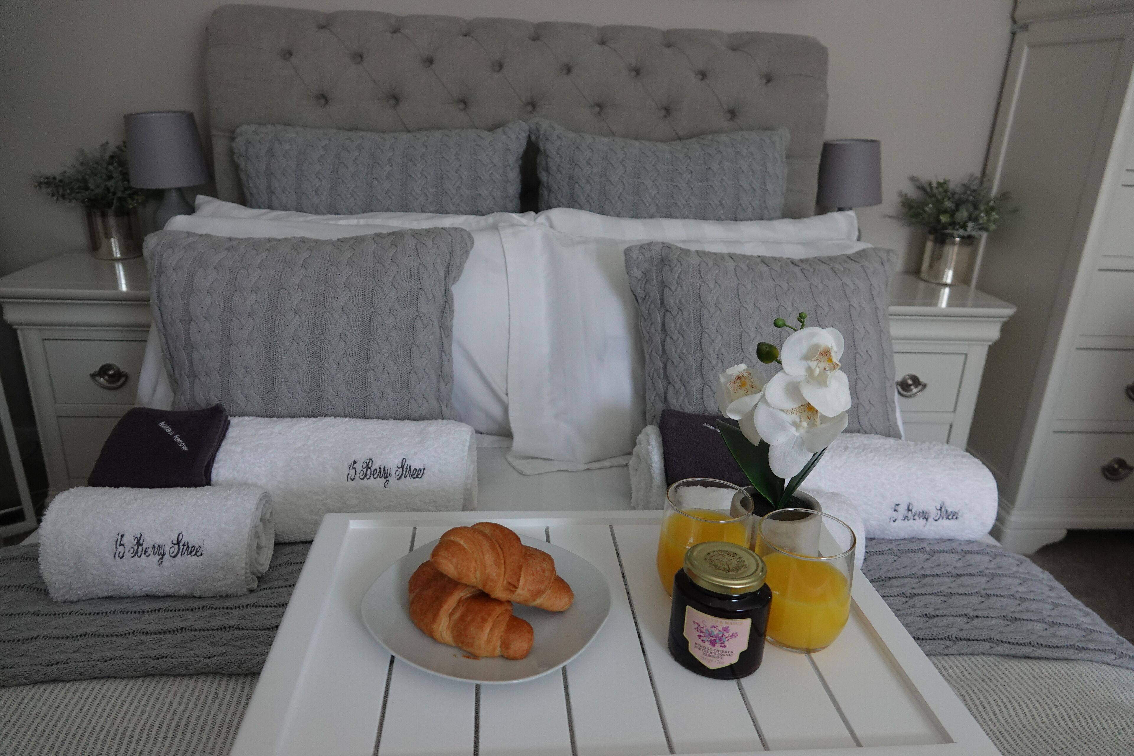 A neatly made bed with grey linens, a tray with croissants, a glass of orange juice, and a jar of jam.