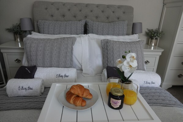 A neatly made bed with grey linens, a tray with croissants, a glass of orange juice, and a jar of jam.