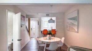 A dining area with a round table, two orange chairs, a pendant light, and a painting of a beach on the wall.