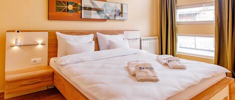 Habitación de hotel con cama de madera, ropa de cama blanca, mesita de noche con lámpara, una obra de arte colgada en la pared y una ventana con cortinas.