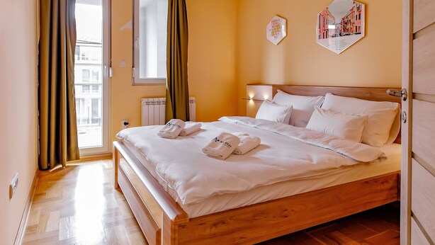 Een net opgemaakt bed met witte lakens en handdoeken, een houten hoofdbord en uitzicht op de stad door een raam met gordijnen.