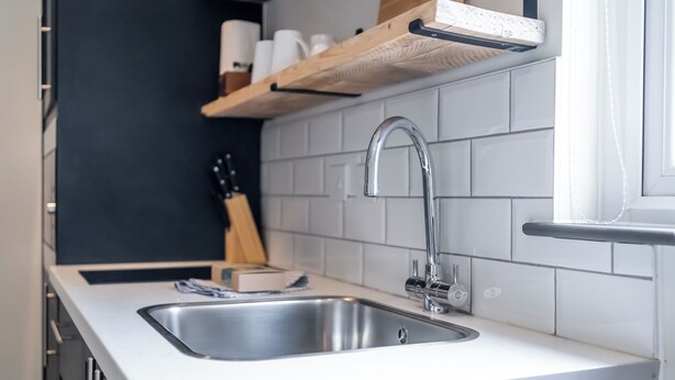 A modern kitchen with a stainless steel sink, white tiles, and wooden shelves above the counter. - Goodstay Lodges by Urban Space (Barry)