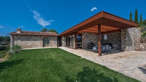 Une maison en pierre avec un toit en tuiles, un patio couvert avec des meubles d’extérieur et une pelouse bien entretenue.