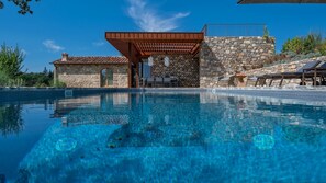 Une piscine d’un bleu éclatant, bordée d’un mur de pierre et surmontée d’une pergola en bois.