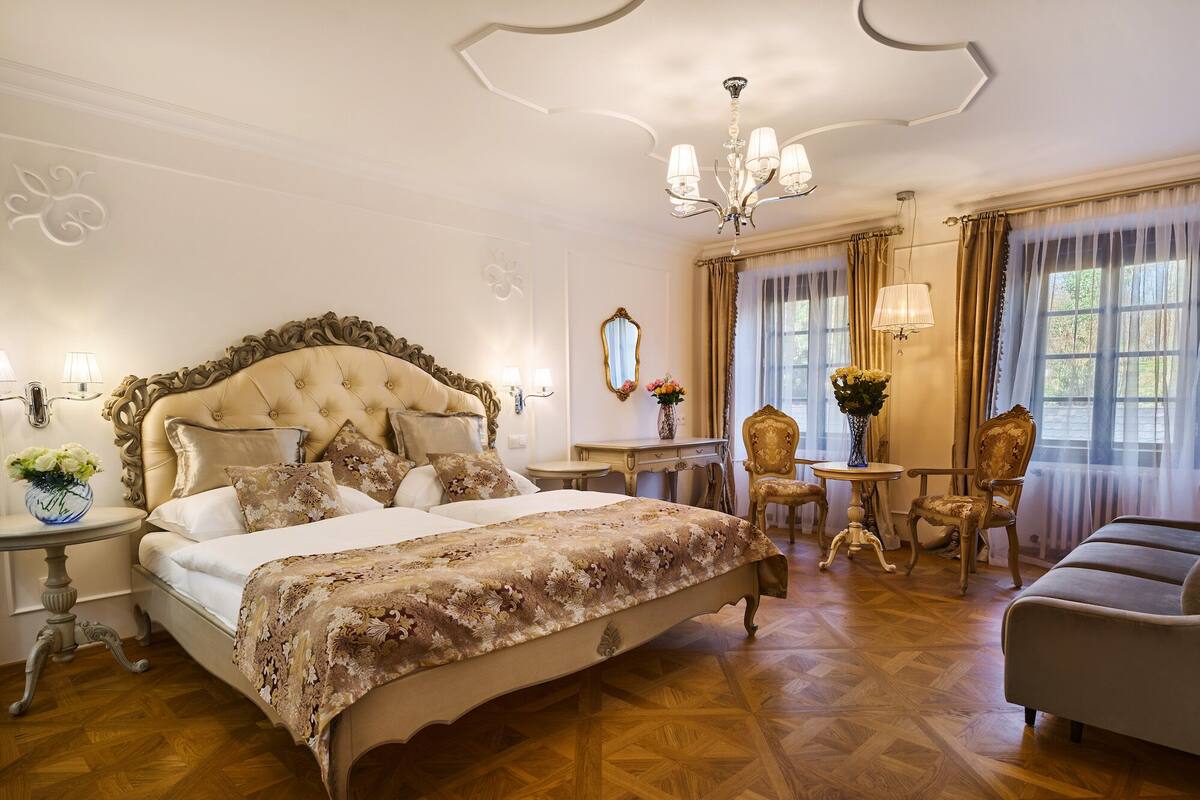 A luxurious bedroom with a large bed, ornate headboard, and a chandelier. There are two chairs and a table with a vase of flowers.