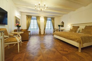Une chambre d’hôtel avec un grand lit, un coin salon comprenant une chaise et une table, et un téléviseur placé sur le mur de gauche.