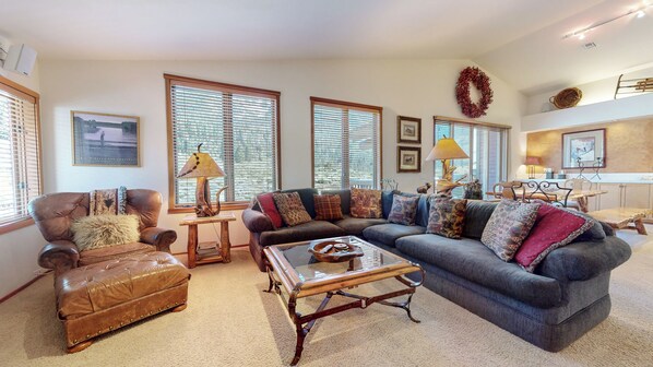 A living room with a brown leather sofa, a blue sofa, and a wooden coffee table. There are large windows with blinds, a fireplace, and a dining area in the background. - Large Family Home. Washer/Dryer, Pool Table & Gas Grill. Mountain Views (Mammoth Lakes)