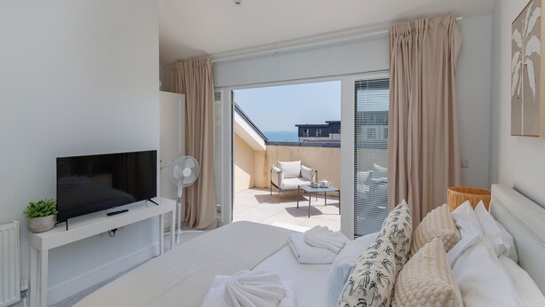 Une chambre dotée d’une télévision, d’un lit avec du linge de lit blanc, d’un balcon offrant une vue sur la mer et d’un fauteuil.