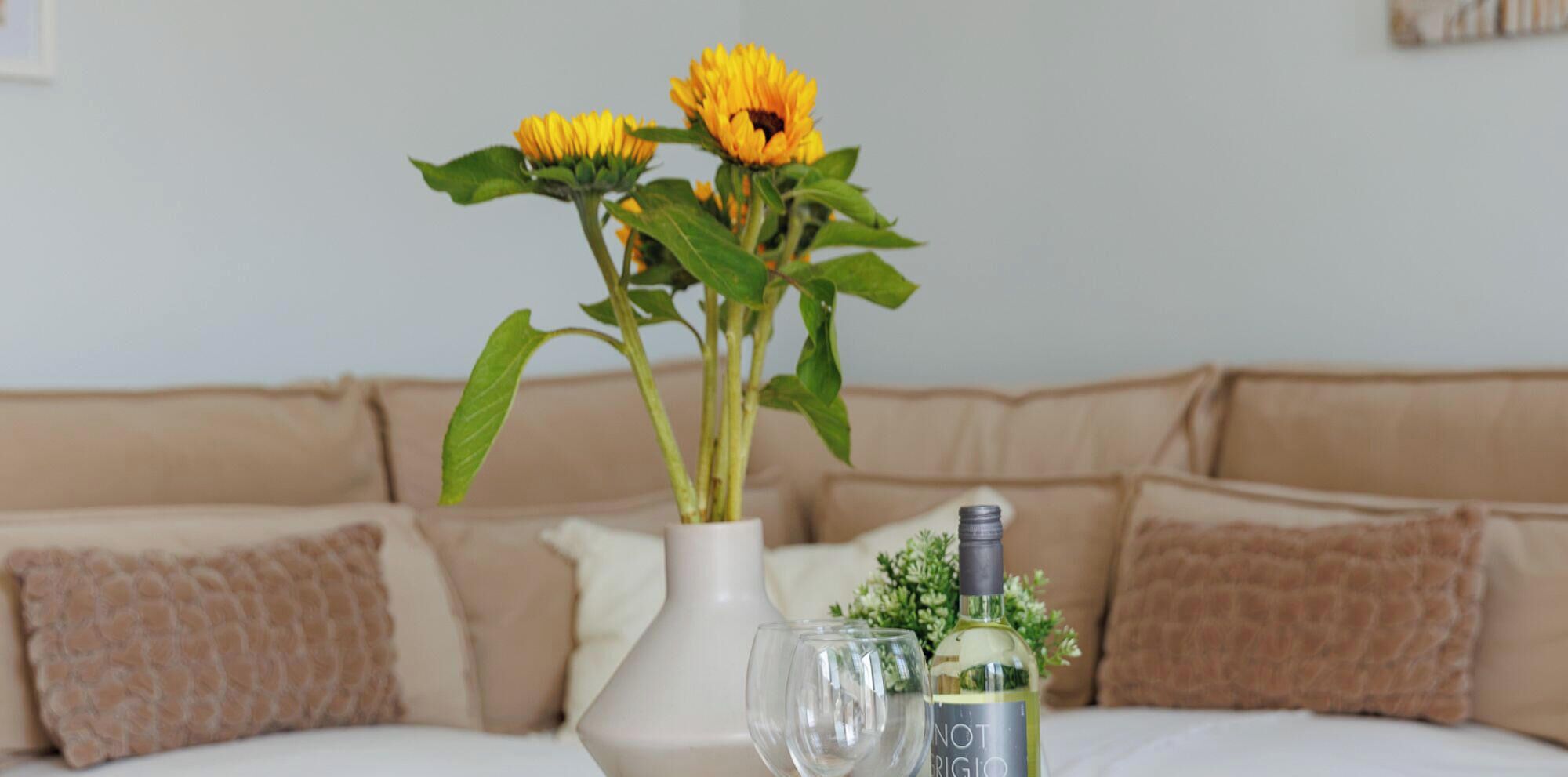 Une pièce dans laquelle on aperçoit un vase blanc contenant des tournesols, une bouteille de vin et un verre posés sur une table.