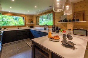 A modern kitchen with a central island, wooden cabinetry, and large windows offering a view of the forest. - Ski Chalet Bonnabelle, Hot tub, garden, parking (Chamonix)