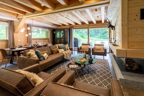 A cozy cabin interior with a wooden ceiling, a dining area, a fireplace, and a large window offering a view of the outdoors.