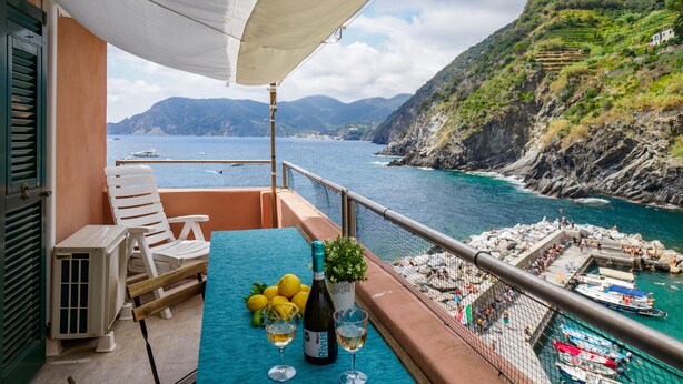 Un balcone con un tavolo apparecchiato con limoni, una bottiglia di vino e bicchieri. Ci sono sedie bianche e una ventola. Si vede un corpo d'acqua e delle barche.