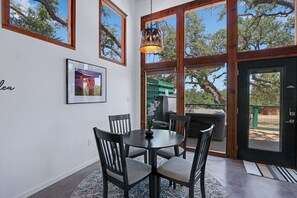 Un espace repas comprenant une table ronde, des chaises, un luminaire suspendu et de grandes fenêtres donnant sur la forêt.