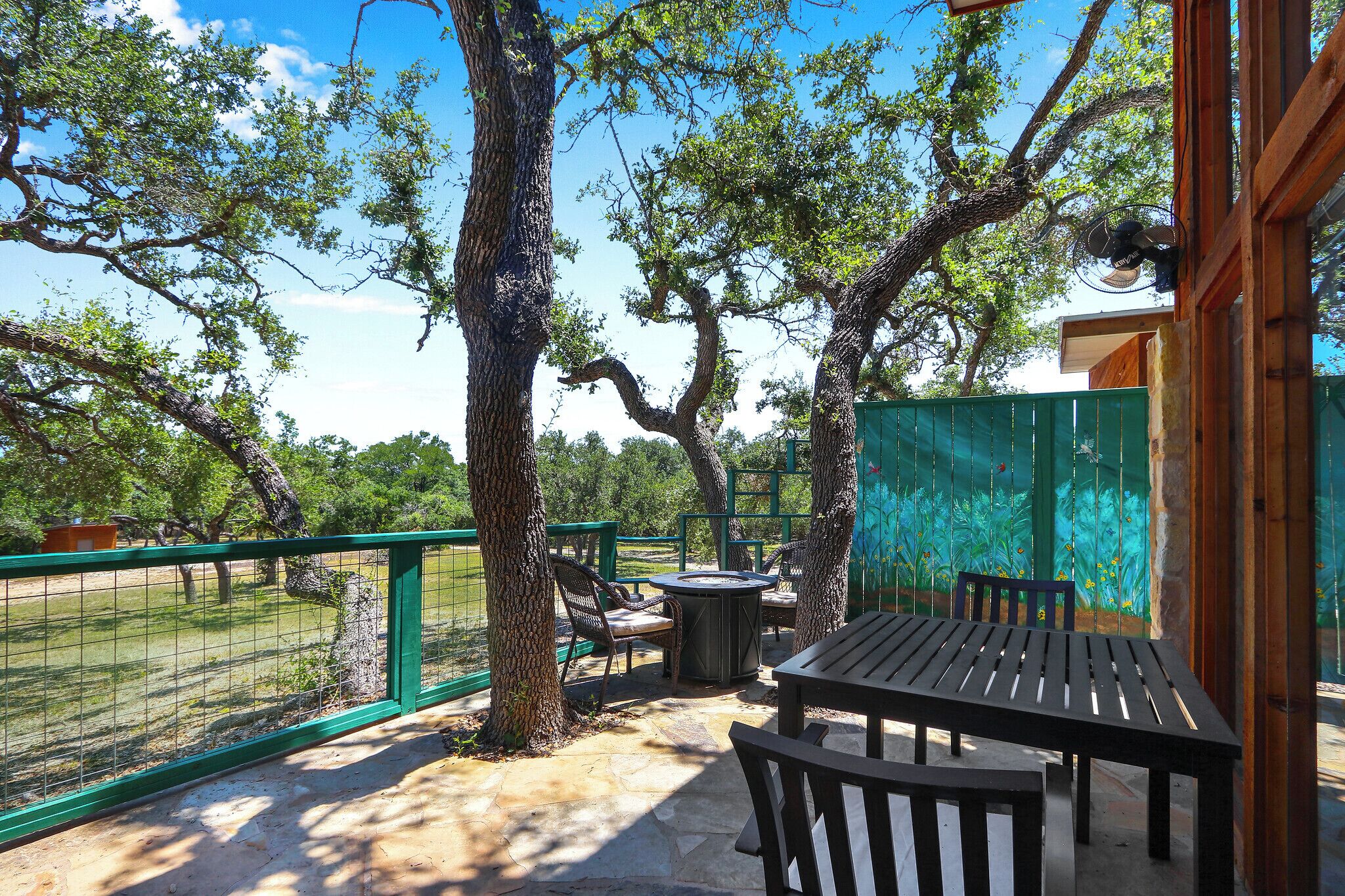 Un área de patio al aire libre con una mesa y sillas, rodeada de árboles y una cerca verde.
