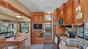 A compact RV kitchen with wooden cabinets, a dining area with a table and chairs, a built-in TV, and a small oven. - Windmill Ranch- Glamping Star RV  I Hot tub, stargazing (Wimberley)