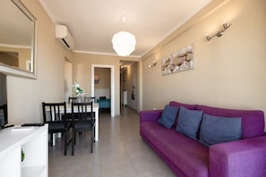 Una sala de estar con un sofá morado, una mesa de comedor con sillas, un televisor y una lámpara de techo.