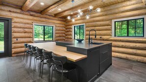 Une cuisine moderne dans une cabane en rondins, avec un îlot central, des tabourets de bar et de grandes fenêtres.