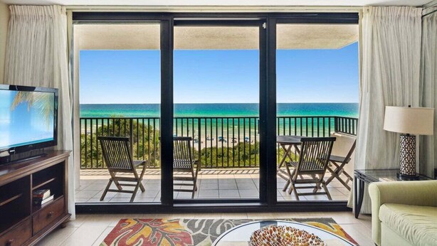 A room with a large sliding glass door leading to a balcony with a view of the ocean. There is a television on the left, a sofa on the right, and a patterned rug on the floor. - Sunset Paradise 308–Corner Unit–Sleeps 8 (Santa Rosa Beach)