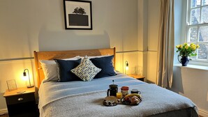 En pænt redt seng med et bakke med morgenmad, et natbord med lampe, og et vindue med gardiner.