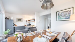Een moderne eetruimte met een houten tafel gedekt voor vier personen, een grijze bank en een keuken op de achtergrond.