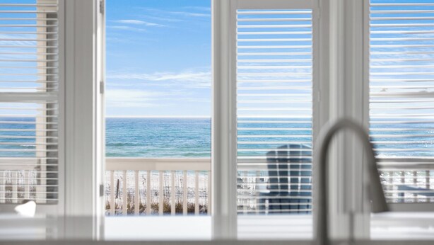 A view from a window with white blinds, overlooking a beach with a clear sky and calm sea. - Gulf/Beachfront Condo | Private Beach (Santa Rosa Beach)