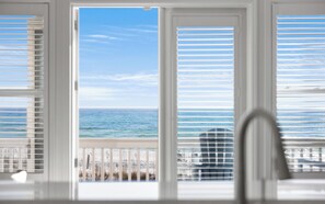 A view from a window with white blinds, overlooking a beach with a clear sky and calm sea.
