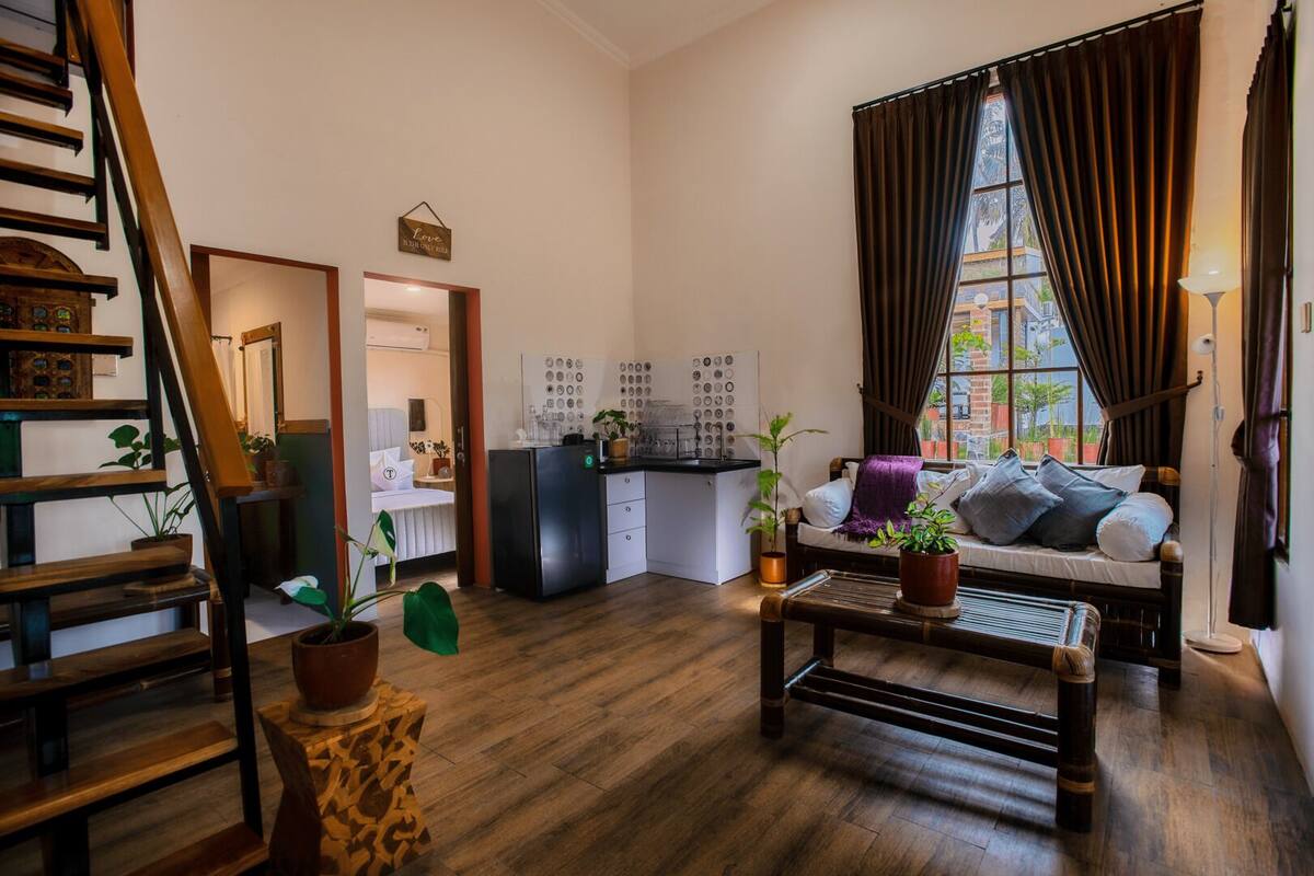 A cozy living room with a wooden staircase, a kitchen area with a refrigerator, and a sofa with cushions.