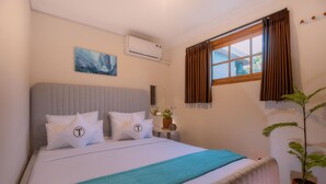 Una cama bien hecha con una colcha azul, un cabecero gris y almohadas con un logo. Se observa una ventana con cortinas, una planta en maceta y un equipo de aire acondicionado en la pared.