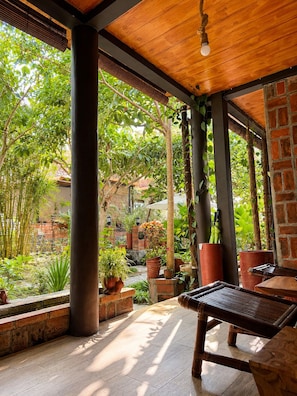 A covered outdoor area with a wooden bench, potted plants, and a view of a garden and buildings in the background. - TZY-NO Cottage Yogyakarta (Pakem)