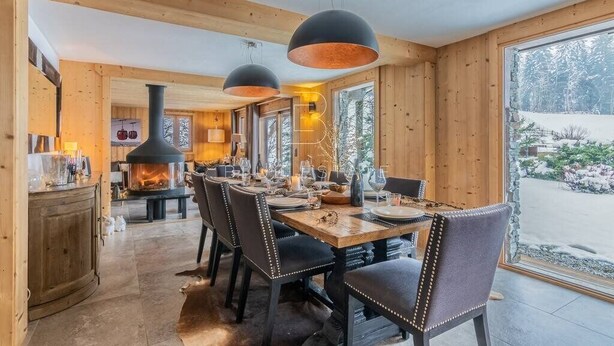 A wooden interior with a dining table set for four, a fireplace, and large windows.