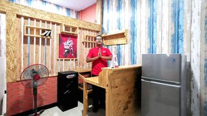 Une cuisine avec un comptoir en bois, un réfrigérateur, un ventilateur et une étagère ornée d’une affiche.