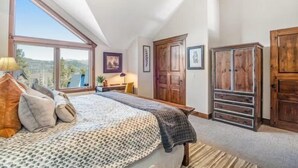 Ein Schlafzimmer mit großem Fenster, einem Bett mit gemusterten Betttextilien, Holzschränken und Blick ins Freie.