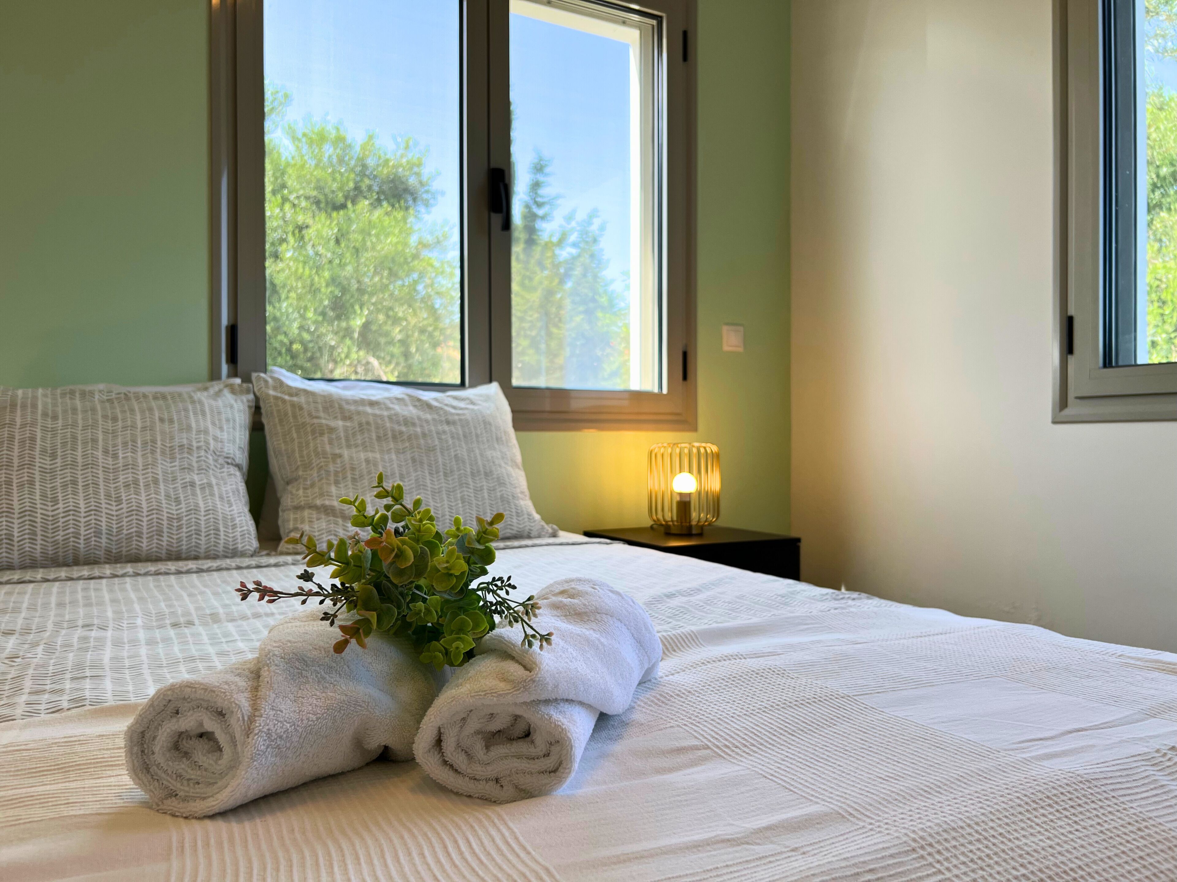A neatly made bed with rolled white towels and a small plant, a bedside lamp, and a window with a view of trees.