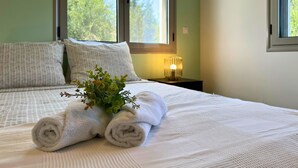 Un lit bien fait, avec des serviettes blanches pliées en rouleau et une petite plante, une lampe de chevet et une fenêtre donnant sur des arbres.